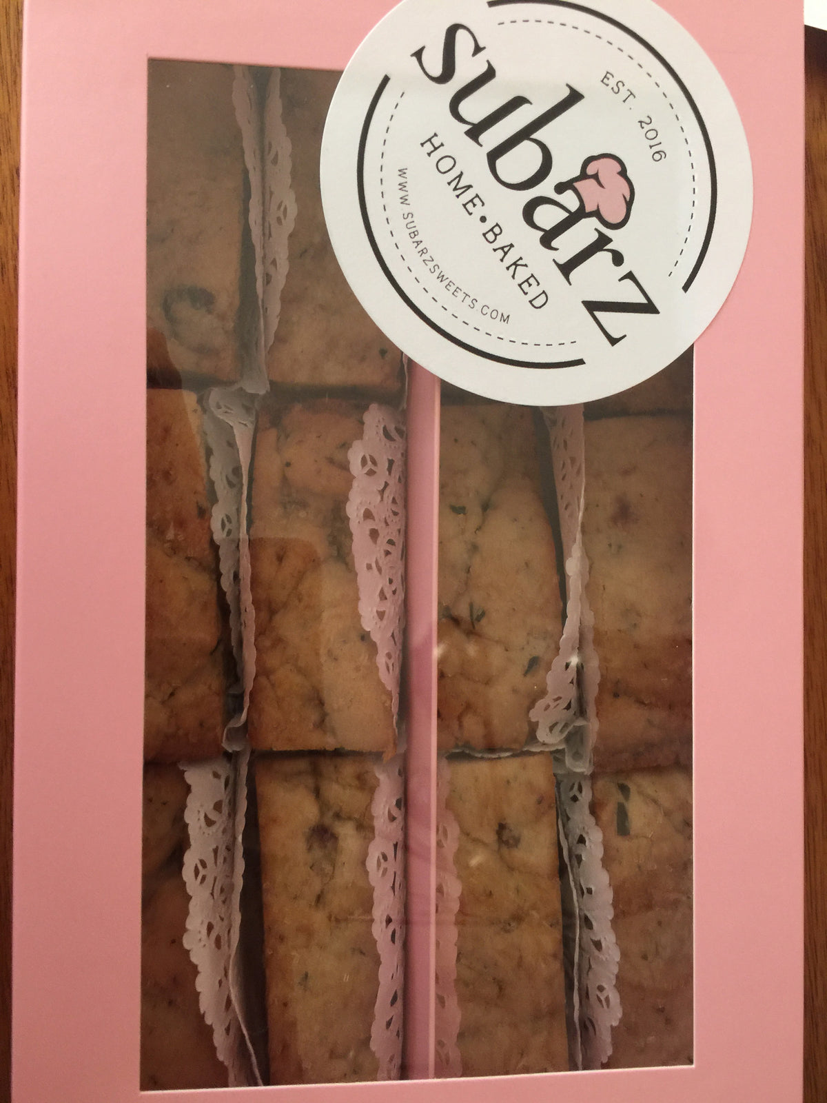 Pink gift box containing one dozen artisanal Subarz, showcased through a windowed display. The golden-brown spiced fig Subarz are arranged in neat rows, accented with delicate white lace paper dividers. The box features the Subarz Home Baked logo, established 2016. The Subarz appear rustic and homemade with visible spices and fig pieces throughout.